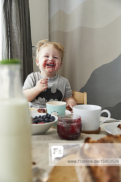 Porträt eines verschmierten kleinen Jungen  der sich am Frühstückstisch vergnügt