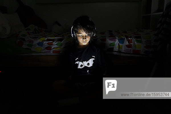 Porträt eines Jungen  der mit Kopfhörer und digitalem Tablett in seinem dunklen Zimmer sitzt
