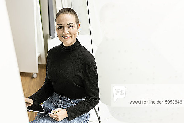 Porträt einer lächelnden jungen Frau  die im Büro am Fenster sitzt und ein Tablett hält