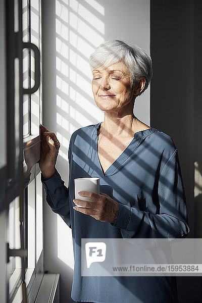 Porträt einer älteren Geschäftsfrau mit Kaffeetasse  die am Fenster steht und das Sonnenlicht genießt
