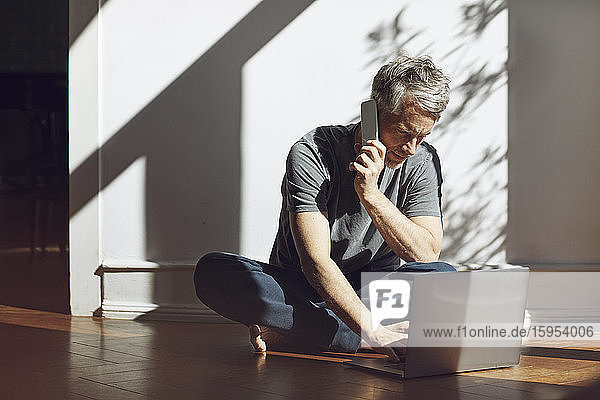 Reifer Mann sitzt zu Hause auf dem Boden und benutzt Laptop und Mobiltelefon