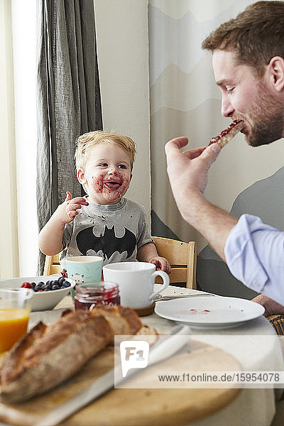 Porträt eines verschmierten kleinen Jungen am Frühstückstisch  der seinen Vater beobachtet  wie er Brot mit Marmelade isst