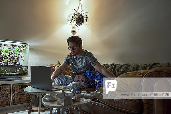 Mann konsultiert Arzt über Laptop  während er bei seiner kranken Tochter sitzt  die zu Hause auf dem Sofa schläft