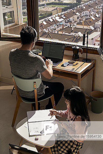 Vater arbeitet am Laptop  während seine Tochter Hausaufgaben macht