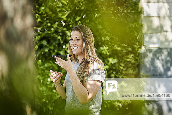 Lächelnde Frau spricht über Smartphone über den Lautsprecher  während sie inmitten von Pflanzen im Hinterhof steht