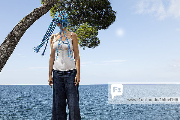 Woman wearing crocheted blue headdress with fringes standing in front of the sea