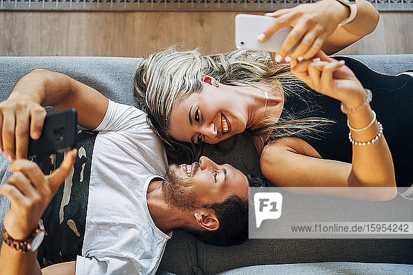 Happy couple lying on the couch taking selfies with smartphones while looking at each other