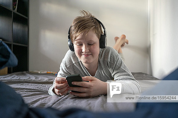 Porträt eines lächelnden Jungen mit Kopfhörern  der auf dem Bett liegt und auf sein Handy schaut