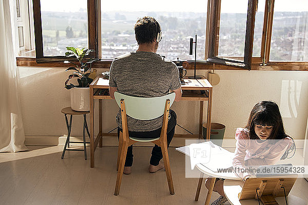 Mädchen macht Hausaufgaben  während ihr Vater im Hintergrund am Laptop arbeitet