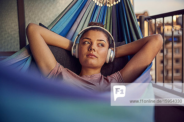 Portrait of young woman lying on hammock on balcony listening music with headphones