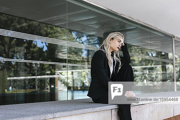 Nachdenkliche junge Geschäftsfrau mit gefärbten blonden Haaren sitzt vor einem Bürogebäude und schaut in die Ferne