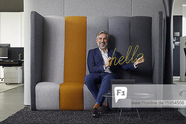 Porträt eines lächelnden reifen Geschäftsmannes  der im Büro auf der Couch sitzt und das Hallo-Schild hält