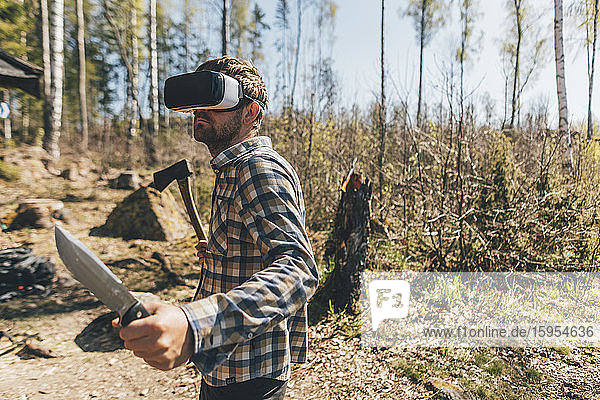 Junger Mann spielt mit VR-Brille im Wald  hält Axt und Messer