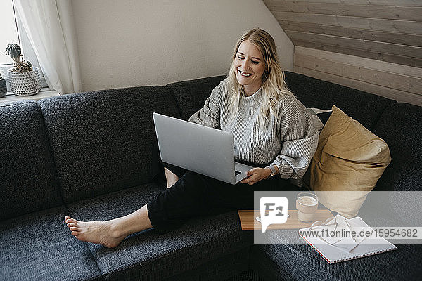 Lächelnder Freiberufler mit Laptop  während er zu Hause mit Getränk und Dokument auf dem Sofa sitzt