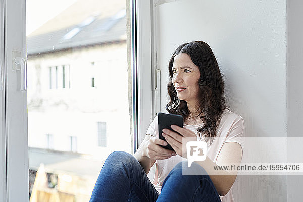 Nachdenkliche Geschäftsfrau mit Smartphone schaut durch das Fenster im Büro