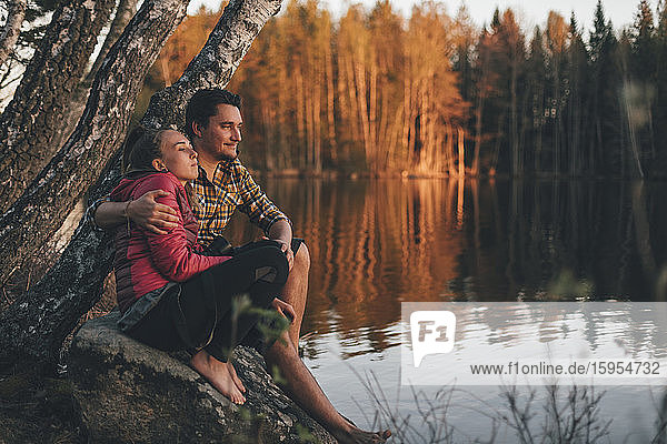 Junges Paar sitzt auf einem Stein am Seeufer und schaut auf den Sonnenuntergang