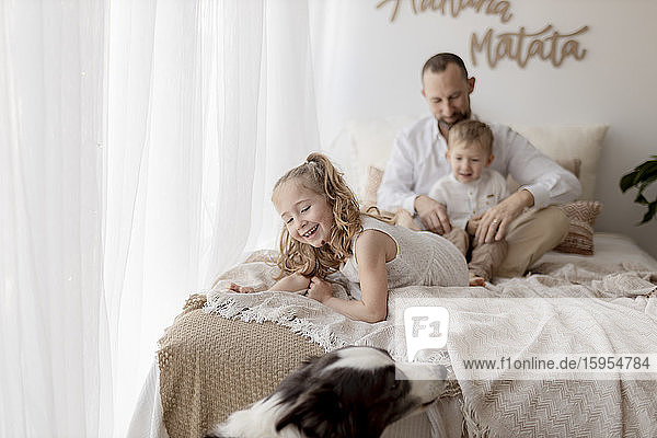 Porträt eines lächelnden kleinen Mädchens im Bett mit Vater und Bruder im Hintergrund