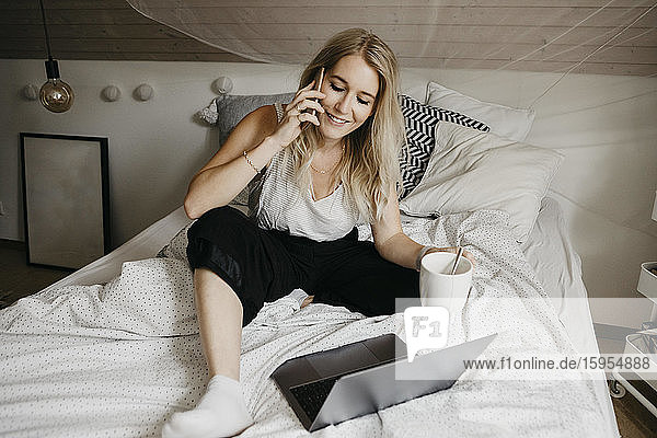 Lächelnde Frau trinkt Kaffee  während sie mit einem Smartphone telefoniert und den Laptop im Schlafzimmer zu Hause benutzt