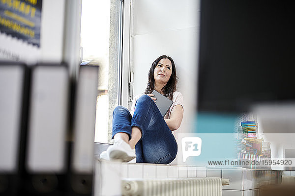 Reife Geschäftsfrau mit digitalem Tablet  die im Büro auf dem Fensterbrett sitzt