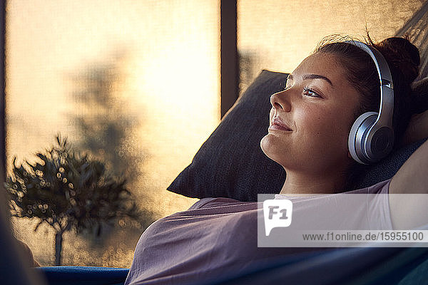 Lächelnde junge Frau liegt auf Hängematte auf dem Balkon und hört Musik mit Kopfhörern