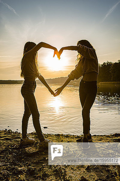 Freunde in voller Länge  die während des Sonnenuntergangs am Seeufer stehend mit den Händen Herzform machen