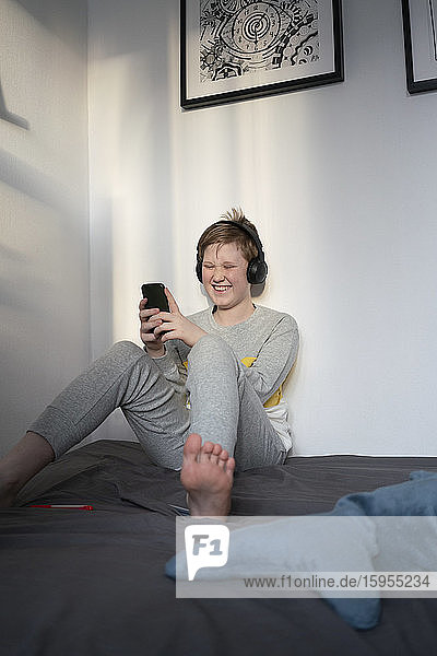 Portrait of laughing boy sitting on bed with headphones looking at cell phone
