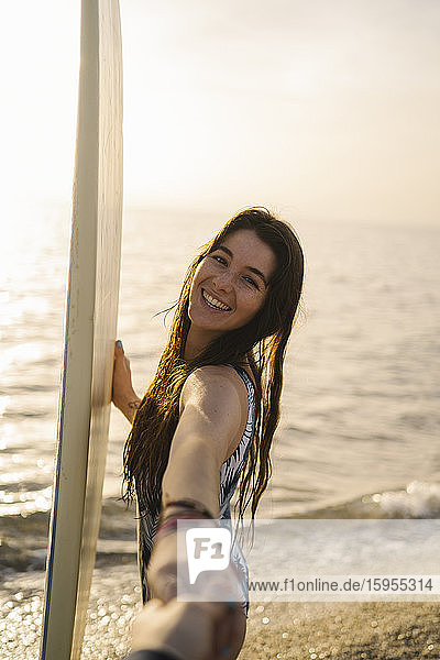 Porträt einer glücklichen jungen Frau mit Surfbrett  die sich am Strand an den Händen hält  Almeria  Spanien