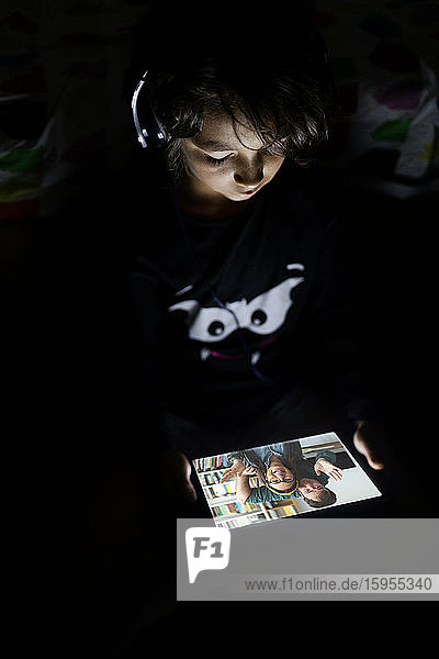 Junge sitzt zu Hause in seinem dunklen Zimmer und benutzt Kopfhörer und digitales Tablet für Video-Chat