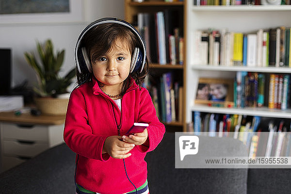 Porträt eines süßen Mädchens  das zu Hause im Wohnzimmer steht und über Kopfhörer Musik hört