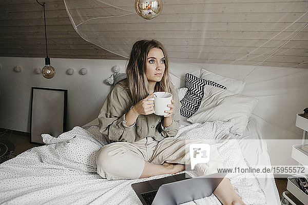 Nachdenkliche Frau mit Laptop  die zu Hause am Bett sitzt und Kaffee trinkt