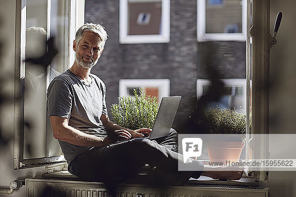 Mature man sitting at the window at home using laptop