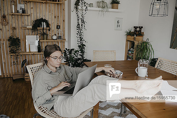 Junge Frau benutzt Laptop  während sie im Haus mit erhobenen Füßen am Tisch sitzt