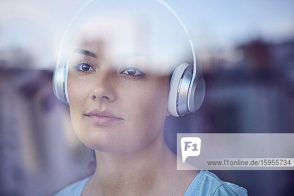 Porträt einer jungen Frau  die mit Kopfhörern Musik hört und aus dem Fenster schaut