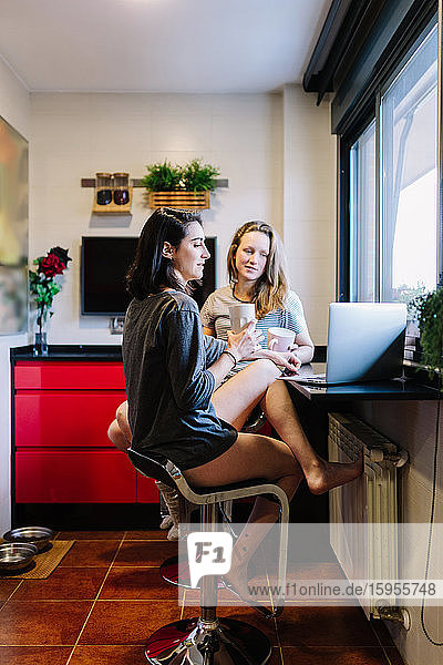 Two women sitting in the kitchen using laptop for video chat