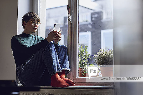 Frau sitzt zu Hause mit Smartphone am Fenster