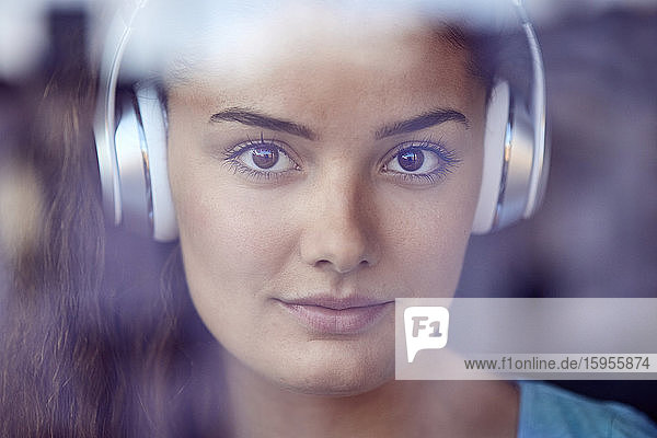 Porträt einer jungen Frau hinter einer Fensterscheibe  die mit Kopfhörern Musik hört