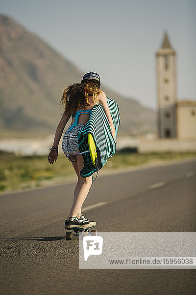 Rückenansicht einer jungen Frau mit einem Surfbrett beim Skateboarden auf einer Asphaltstrasse  Almeria  Spanien