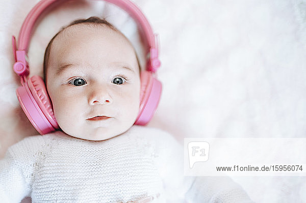 Porträt eines kleinen Mädchens mit übergrossen rosa Kopfhörern auf einer weissen Decke liegend