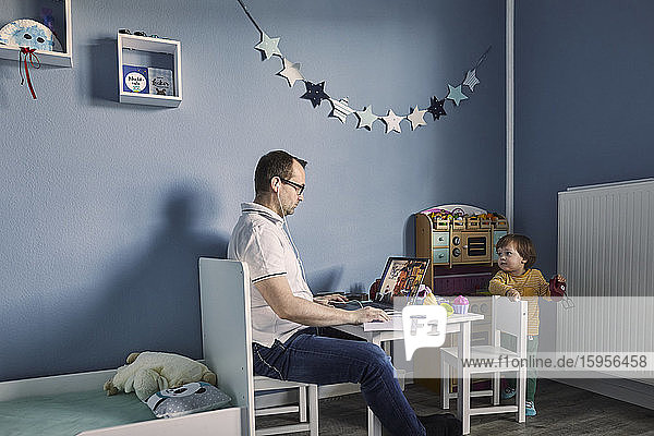 Mann arbeitet und benutzt Laptop im Kinderzimmer mit gleichzeitiger Kinderbetreuung