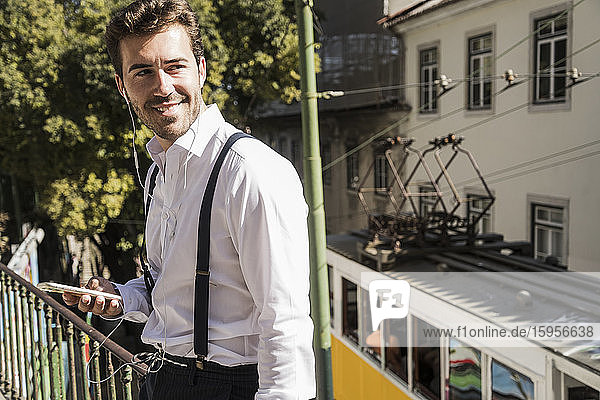 Lächelnder junger Mann mit Smartphone und Ohrstöpseln in der Stadt  Lissabon  Portugal