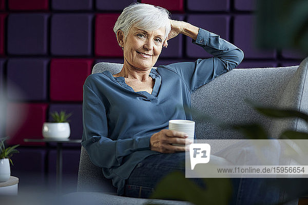 Porträt einer lächelnden älteren Geschäftsfrau  die auf einem Sessel mit Kaffeetasse sitzt