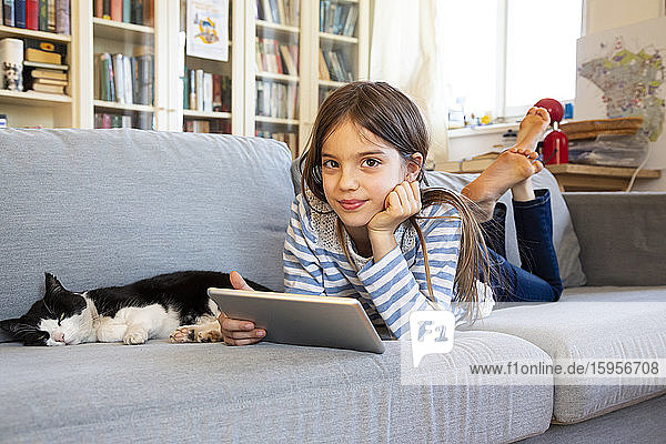 Porträt eines lächelnden Mädchens auf einer Couch liegend mit Katze und digitalem Tablett
