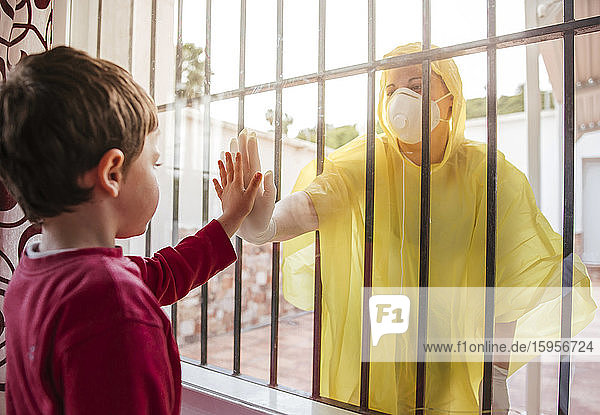 Junge  der die Hand der Mutter berührt und Schutzkleidung hinter der Fensterscheibe trägt