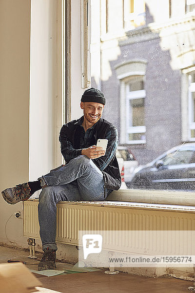 Mann renoviert Ladenstandort  sitzt auf der Fensterbank  benutzt Smartphone