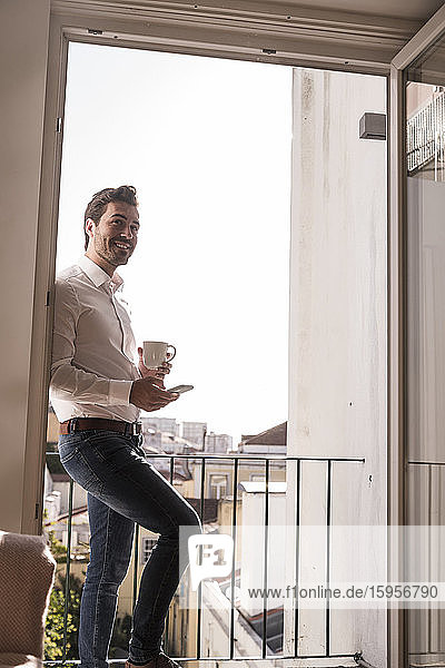 Junger Mann mit Handy und Kaffeetasse auf einem Balkon