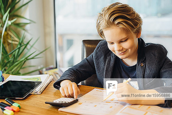 Lächelnder Junge am Schreibtisch sitzend mit Arbeitsbuch und Taschenrechner