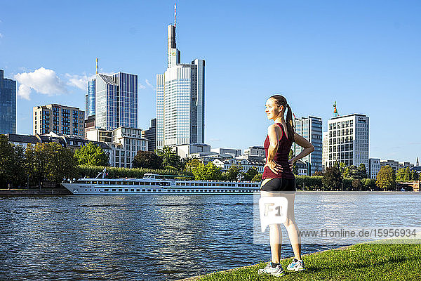 Deutschland  Hessen  Frankfurt  Junge Frau in Sportkleidung am Mainufer stehend mit Stadtsilhouette im Hintergrund