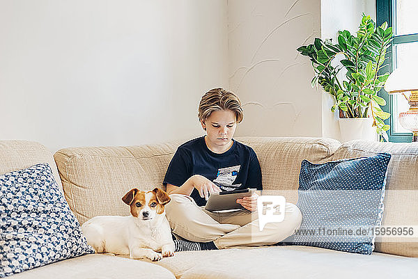 Junge mit Hund und Tablett auf der Couch sitzend im Wohnzimmer zu Hause