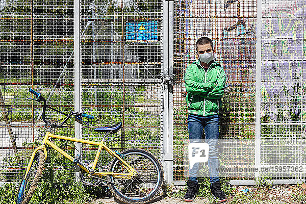 Porträt eines vorpubertären Jungen  der eine Gesichtsmaske trägt  während er mit verschränkten Armen mit dem Fahrrad gegen den Zaun steht