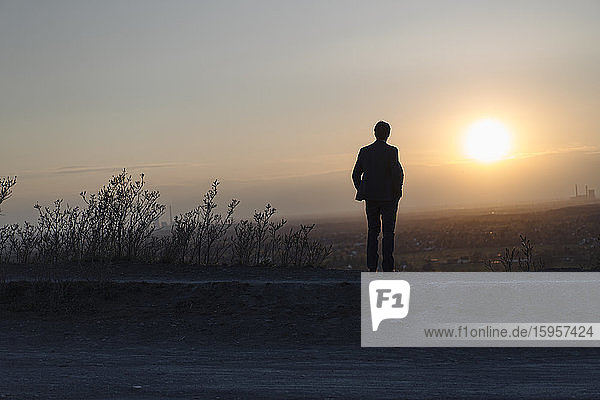 Ein reifer Geschäftsmann  der bei Sonnenuntergang auf einer stillgelegten Minenhalde steht und auf die Aussicht schaut